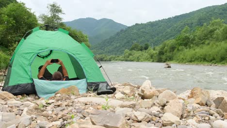 a tourist rests in a green tent on the bank of a mountain river looking at the phone a tourist rests in a green tent on the bank of a mountain river looking at the phone rest in the fresh air in the middle of the mountains