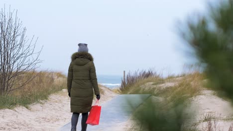 mujer joven caminando por la playa en un día nublado de invierno, bolsa roja en la mano derecha, pino desenfocado en primer plano, tiro medio de mano