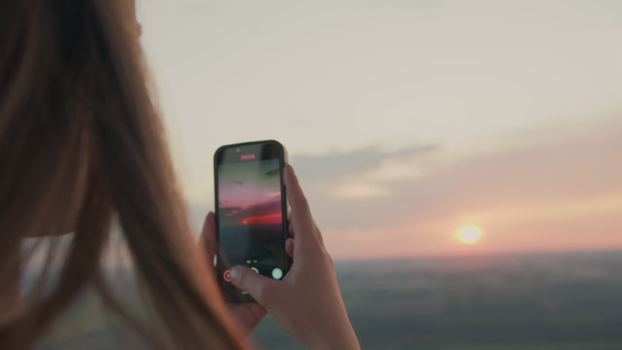 Rear view of woman holding smartphone to take photo of glowing sunrise horizon while floating above scenic farmland, capturing peaceful moment with soft light and serene sky