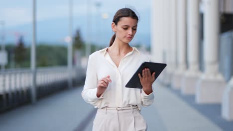 Young-attractive-businesswoman-walks-along-modern-building-with-tablet