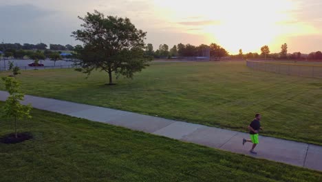 Stetige-Drohnenaufnahmen-Eines-Mannes,-Der-Durch-Den-Park-Rennt,-Mit-Sonnenaufgang-Im-Hintergrund