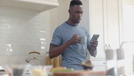 Happy-african-american-man-drinking-coffee-and-using-smartphone-in-sunny-kitchen,-slow-motion