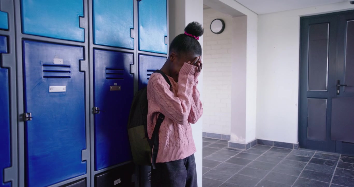 African preteen standing in pink sweater by blue school lockers, two peers pointing, covering face