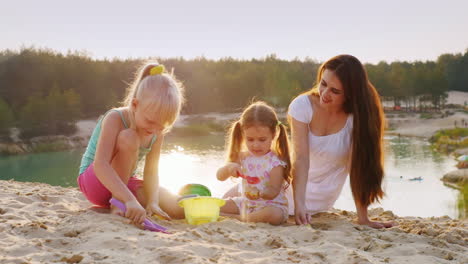 young mother plays in the sand with two daughters at sunset on the background of the beautiful lake