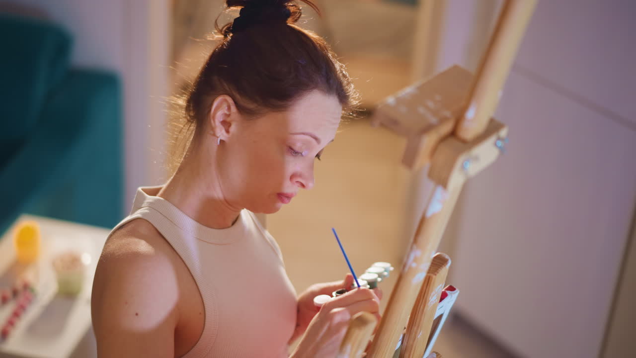 Woman Creating Art With Careful Strokes, Caucasian Artist Painting Thoughtfully At Studio Easel, Woman With Light Hair Meticulously Painting At Easel Surrounded By Art Supplies Under Warm Lighting