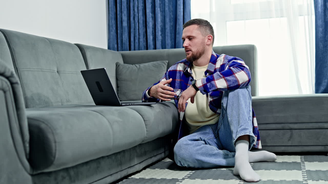 Man sits on the floor with a laptop in a living room while wearing socks and a flannel shirt during the daytime near a couch and a window