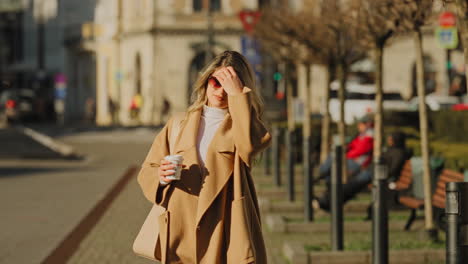 movimiento lento de una joven hermosa mujer rubia caminando con confianza y sonriendo, sosteniendo una taza de café para llevar, en una mañana soleada