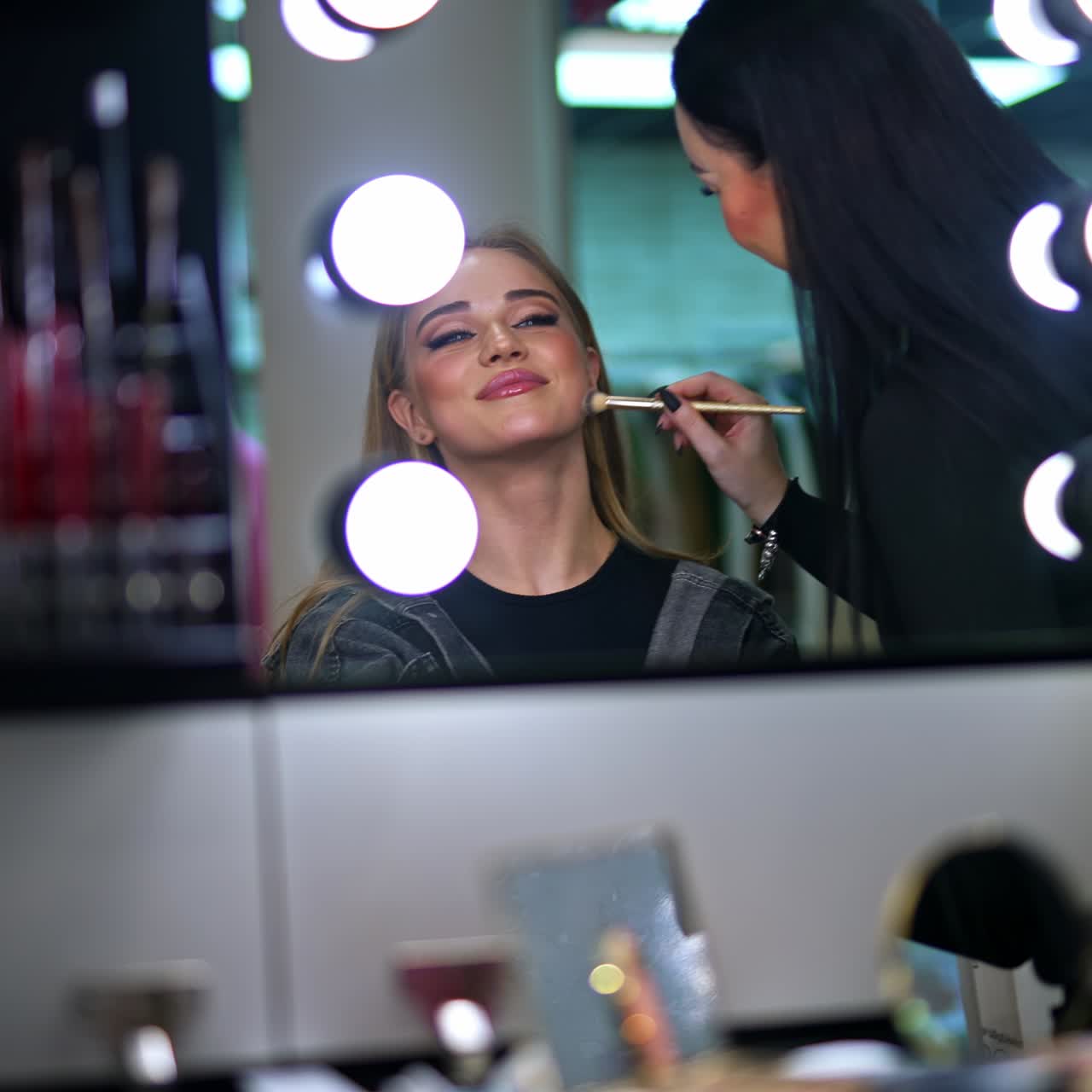 Young blond girl with bright make-up sitting in the chair looking in the mirror with lamps. Makeup artist uses brush on a lady's smiling face