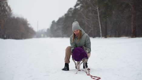 woman crouching in snowy park giving loving attention to dog wearing purple coat during winter walk, gently caressing its face, surrounded by falling snow, expressing warmth, care, and joyful companionship