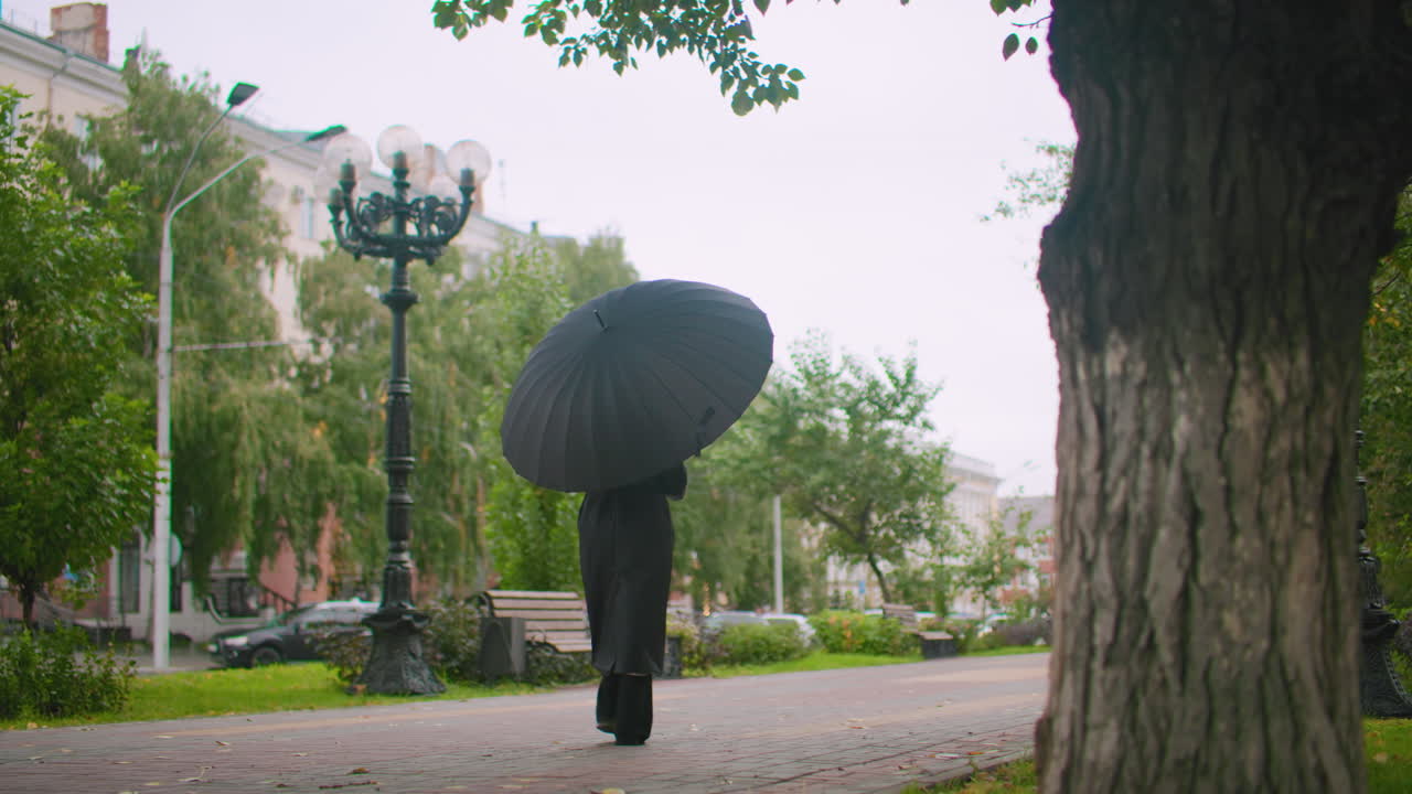 Person wearing long black coat holding open black umbrella walks through city park on cloudy day, surrounded by trees, benches, buildings, and cars, suggesting rainy urban lifestyle scene