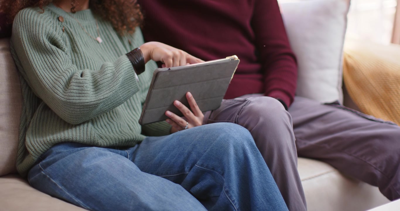 Par sitter i sofaen, kvinne trykker på nettbrettmappen mens mannen lener seg inn og ser på bilder.