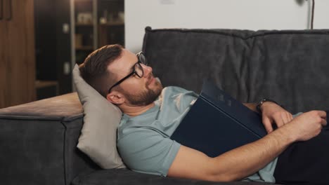 an adult man quickly falls asleep while lying on the couch reading a book