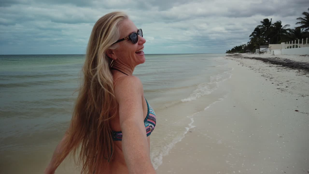 Closeup rear shot of playful blonde mature woman in sunglasses and bikini running along a beach and looking back at the camera with arm back reaching out to someone