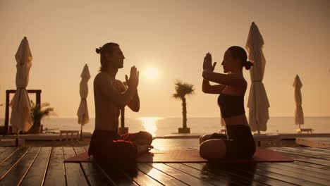 una chica morena en una camiseta negra y un tipo con un torso desnudo están meditando en una alfombra roja en una playa soleada durante un amanecer dorado