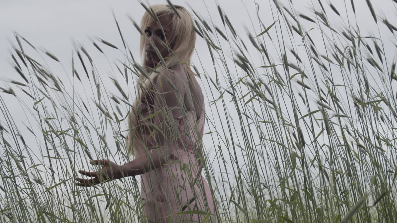 joven negra en vestido rosa, con el cabello rubio largo; de pie en un campo de hierba alta. su espalda está a la cámara mientras corre con las manos a lo largo de la hierba alta balanceándose. 60 fps cámara lenta, cámara de mano.