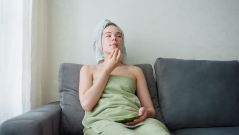 girl sitting on couch with crossed legs and stretched-out feet, wrapped in towel, enjoying quiet moment in calm indoor environment filled with natural light through window, reflecting comfort