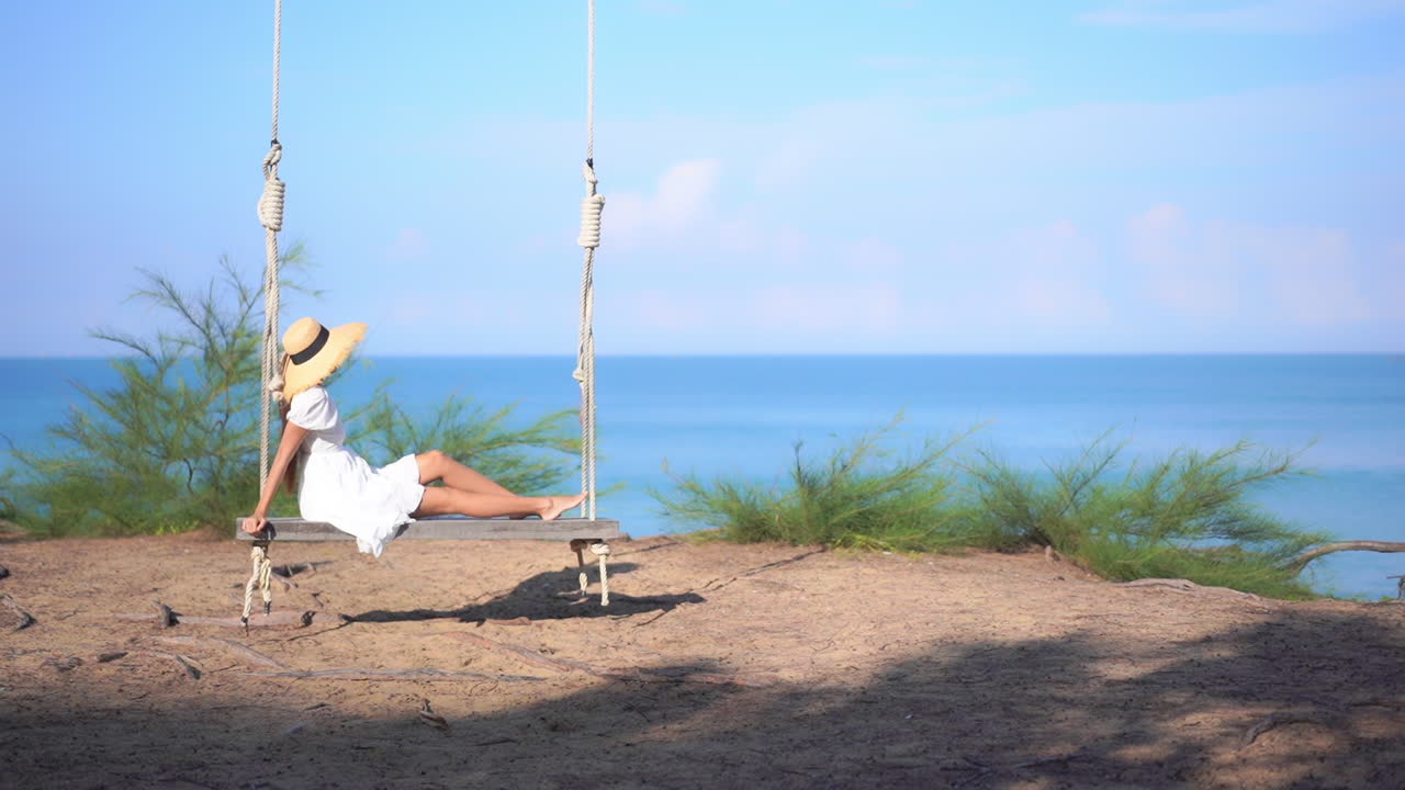 mujer irreconocible en vestido de verano y sombrero balanceándose en un gran columpio de banco con cuerdas en un increíble fondo con vistas al mar en tailandia