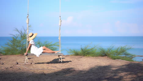 unrecognizable woman in sundress and hat swinging on a big bench swing with ropes on amazing sea view background in thailand