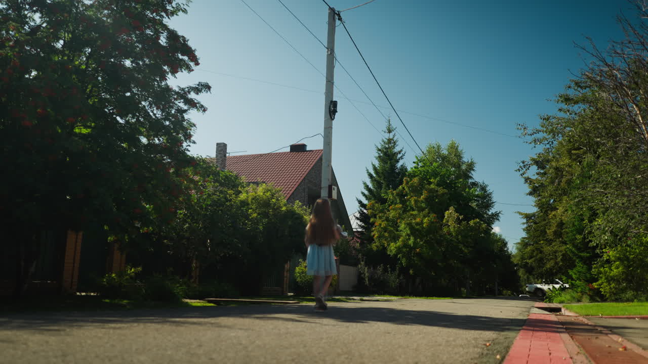 Back view of child in gown walking peacefully along residential street under clear sky, surrounded by lush green trees and houses, casting shadow on pavement during sunny afternoon stroll