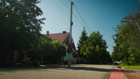 back view of child in gown walking peacefully along residential street under clear sky, surrounded by lush green trees and houses, casting shadow on pavement during sunny afternoon stroll
