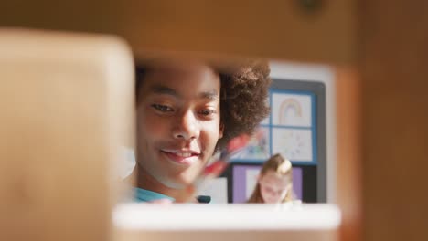 video of happy african american schoolboy painting in art class, seen through easel, copy space