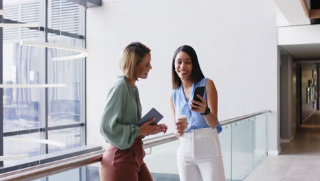 holding smartphone, two businesswomen laughing and chatting during office break
