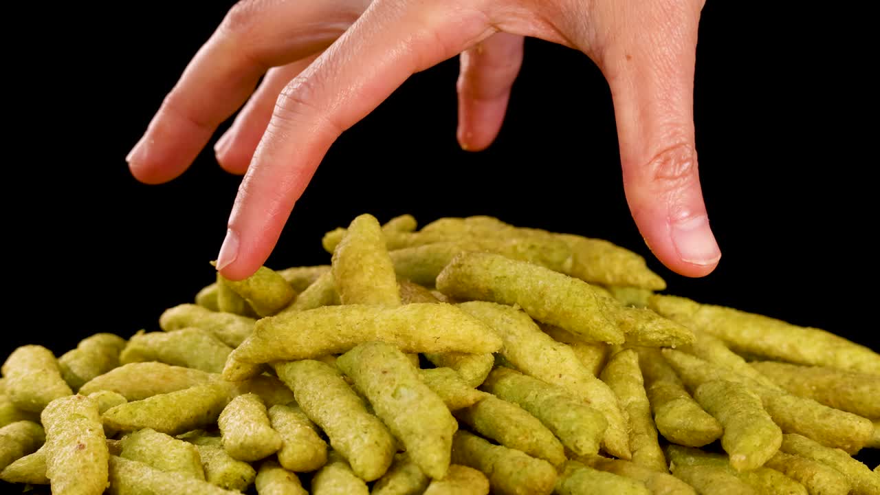 Human hand selects and lifts green pea puffed snacks under bright studio lighting, black background