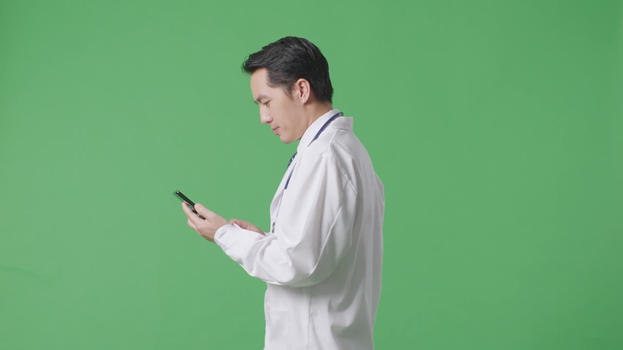 병원에서 녹색 화면 배경에서 는 동안 스마트폰을 사용하여 스테토스코프를 가진 아시아 남성 의사의 측면 시각