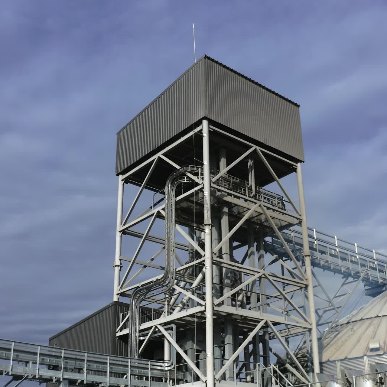 High metal construction standing next to the elevator tanks. Looking at the tops of modern granary buildings at the backdrop of grey cloudy sky