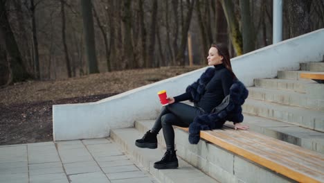 sexy young woman with a cup of coffee outdoors. beautiful lady sitting alone on the bench in the park in cold season and drink tea. slow motion.