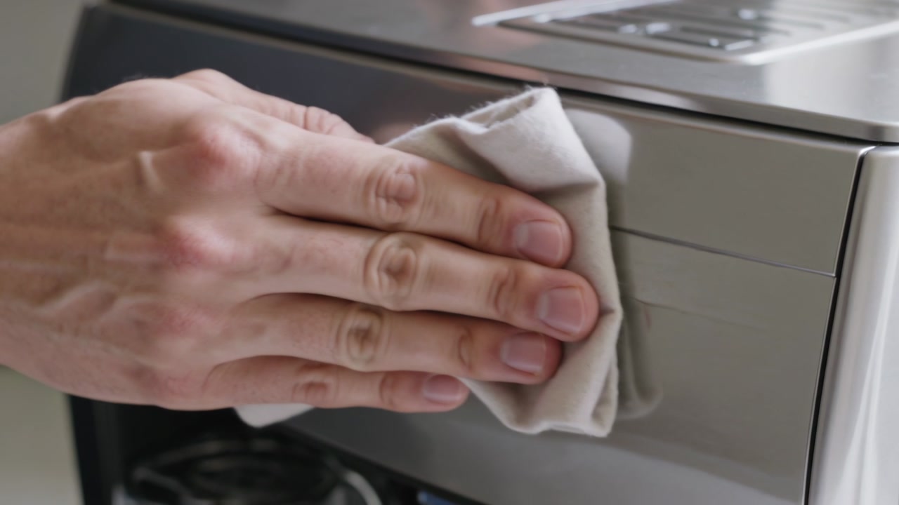Wiping hand coming from right, sliding cloth across steel coffee maker in kitchen, removing smudges