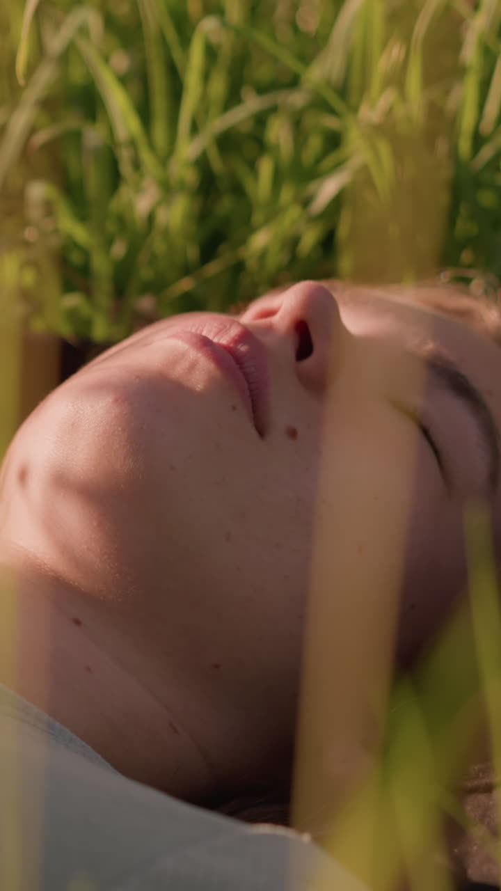 primer plano de una mujer joven acostada en un campo cubierto de hierba, mirando pensativamente hacia arriba, inmersa en la naturaleza, rodeada de hierba alta y iluminada por el sol que enmarca suavemente su rostro