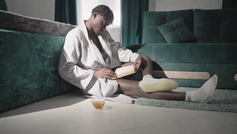 man in white robe sitting on rug, reading book and enjoying tea in stylish, modern bedroom. morning light shines softly on floor, creating peaceful, cozy atmosphere for a relaxing start to the day