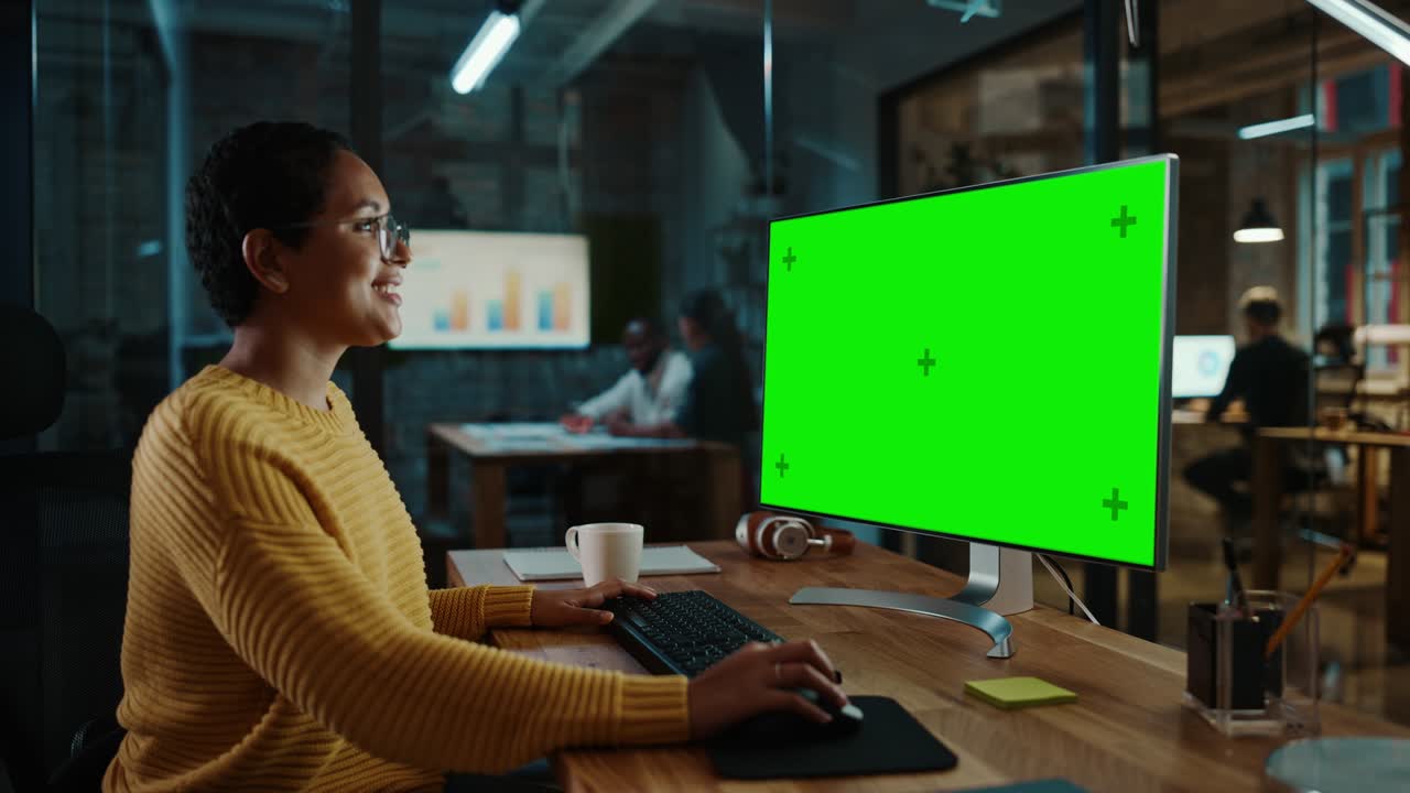 giovane specialista multietnico che lavora su un computer desktop con schermo verde in un ufficio creativo. bellissima manager femminile diversificata con i capelli corti e gli occhiali indossa un maglione giallo.
