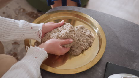high angle view of therapist carrying golden tray with soil mold while approaching sofa where visitor sits and rubs soil during interactive therapy session in cozy room with soft natural lighting