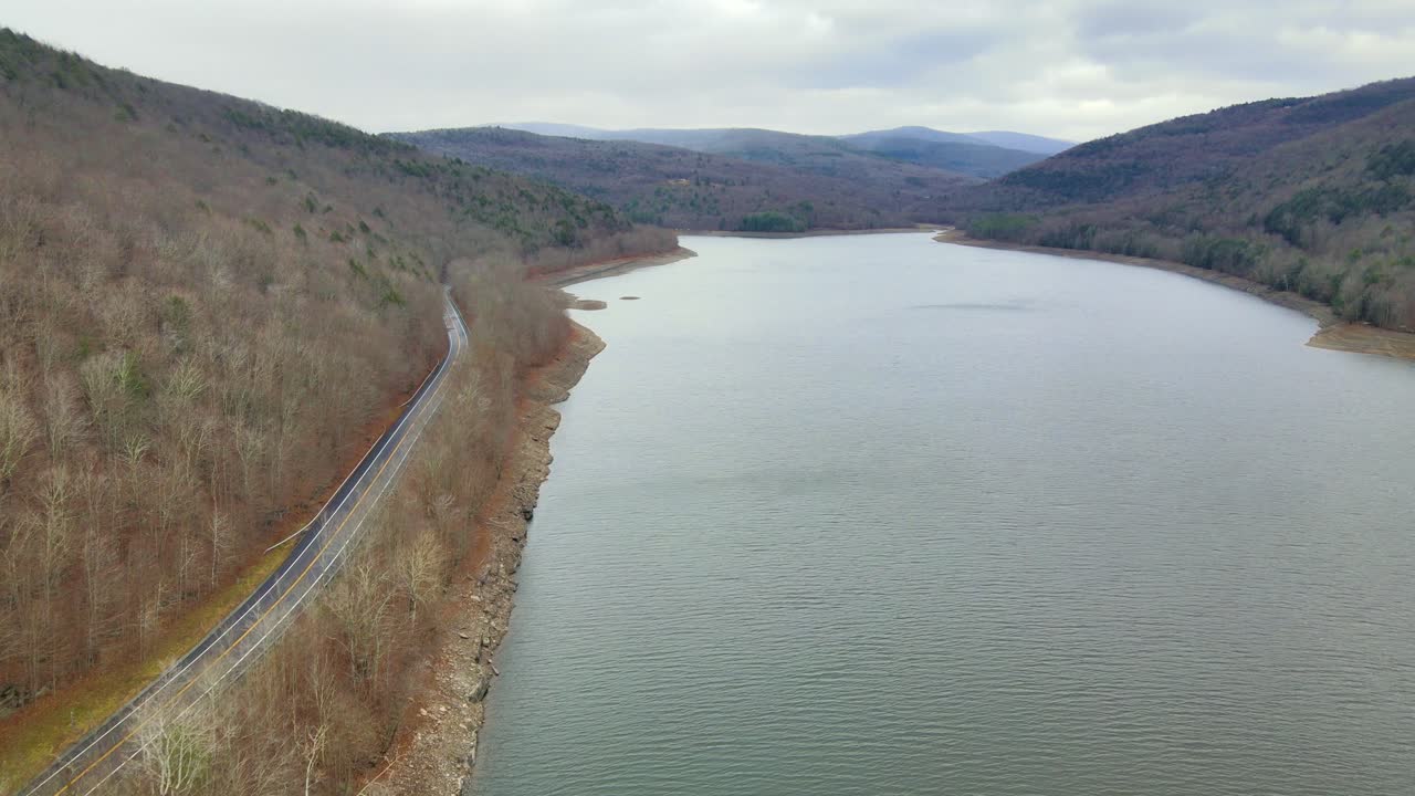 drone volando hacia adelante sobre un lago de montaña junto a una carretera de montaña a principios de invierno, sobre un valle de montaña