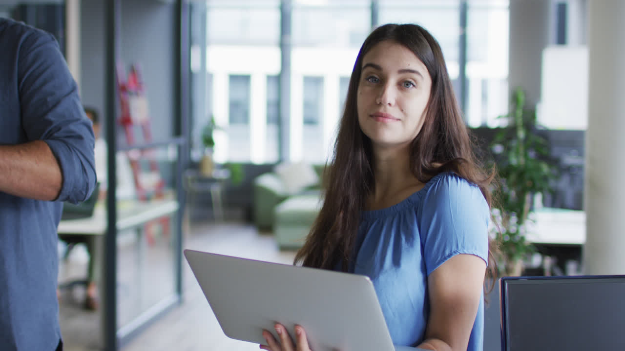 retrato de una mujer de negocios caucásica de pie en la oficina usando una computadora portátil, mirando a la cámara y sonriendo