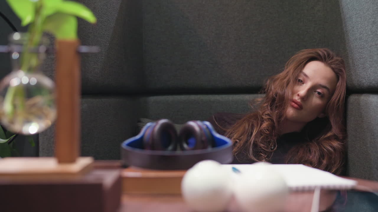 Lady with curly brown hair rests head against cushioned chair wall, eyes tired and face weary, as closed headset, open notebook, and plant decoration sit on table in front, suggesting quiet fatigue