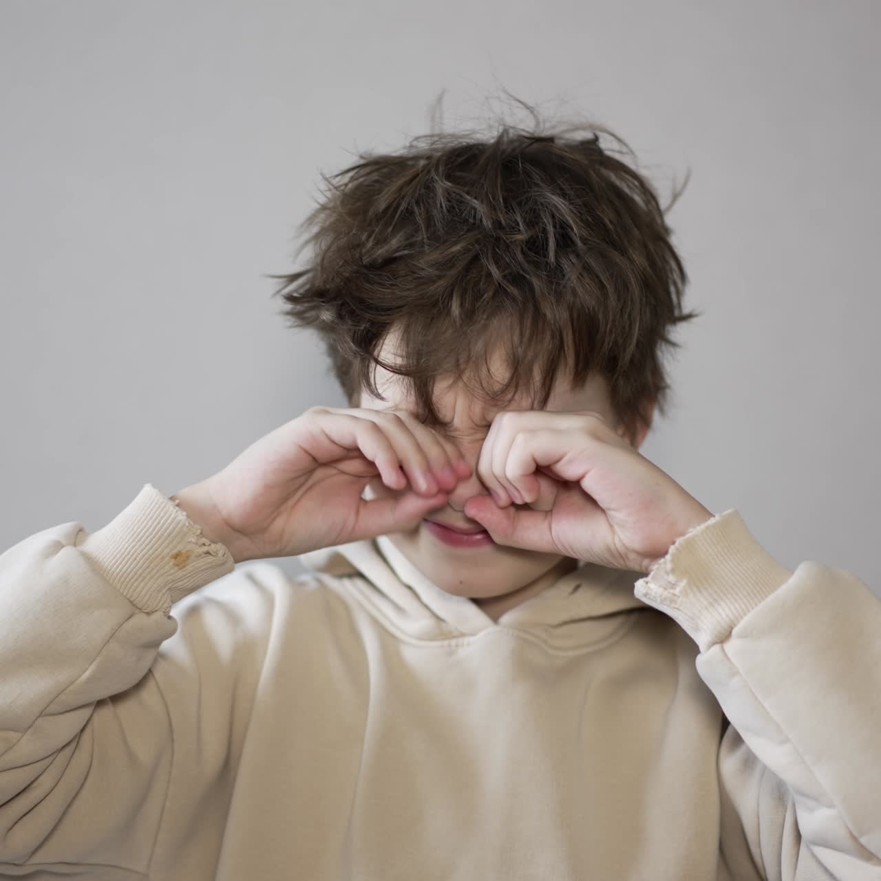 Kid wearing beige hoodie rubbing his eyes and messing his hair. Teenage boy in a bad mood