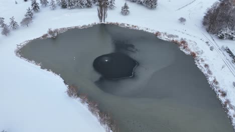 Luftaufnahme-Eines-öffentlichen-Teichs-Mit-Einem-Brunnen-In-Der-Mitte-Und-Schnee,-Der-Die-Küste-Bedeckt