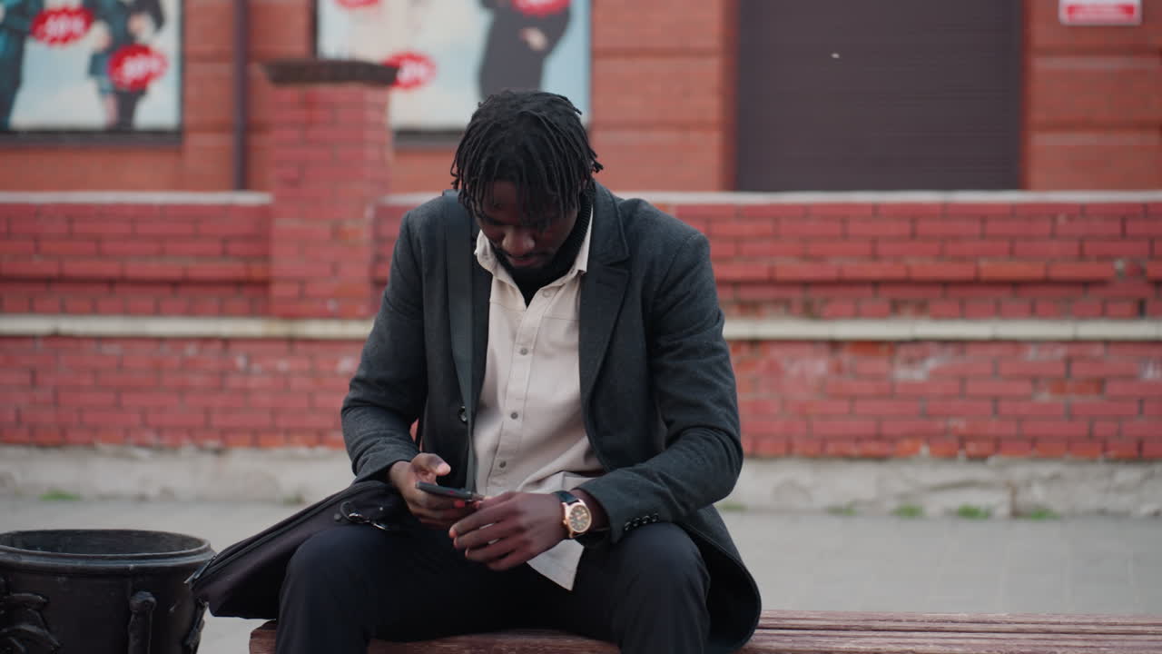 gentle Man sitting on bench using phone with street advertising posters in background, dressed in light shirt, portraying urban lifestyle, focus, and digital engagement in modern city environment