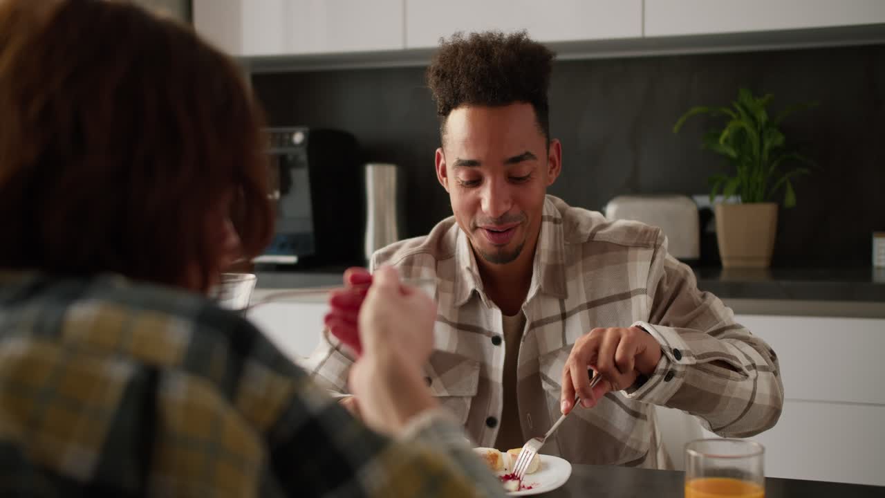 Over the shoulder a happy young Black-skinned brunette man in a cream plaid shirt eats cheesecakes with jam and communicates with his young adult girlfriend in a plaid shirt during their breakfast together in a modern kitchen in an apartment