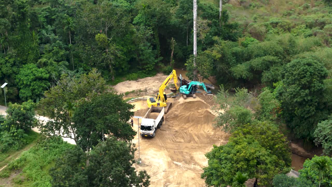Aerial view of construction work in progress on Koh Samui, Thailand. Heavy machinery like excavators and cranes are actively moving earth in a lush, green environment.