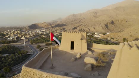 zdjęcie lotnicze o zachodzie słońca z fortu dhayah w ras al-khaimah w zjednoczonych arabskich emiratach, z flagą emiratów w pełnym fokusie, ujawniając miasto poniżej