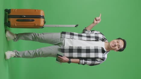 full body of asian male traveler with luggage and passport smiling and pointing to side while standing in the green screen background studio