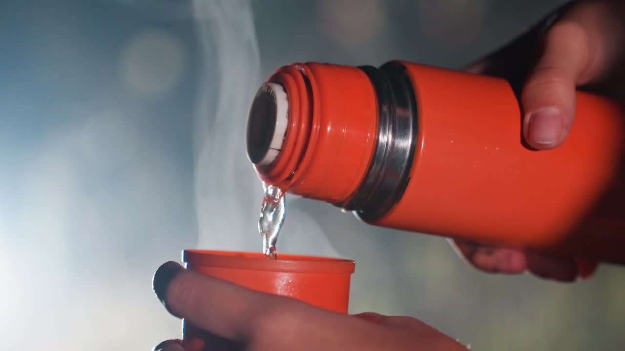 Un thermos arancione fumante versa una bevanda calda, mani a coppa che sorreggono una tazza piccola durante l'osservazione notturna delle stelle vicino al telescopio, luci bokeh soffuse, alito freddo visibile, atmosfera da campeggio, atmosfera da osservazione scientifica, primo piano