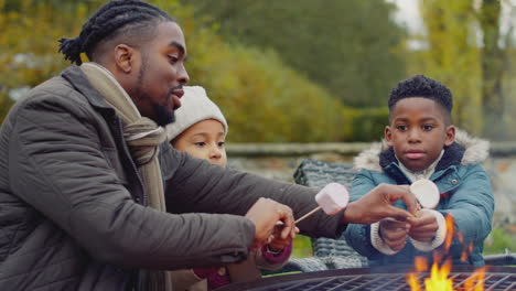 father helping children as family toast marshmallows sitting around firepit or barbeque in garden at home - shot in slow motion