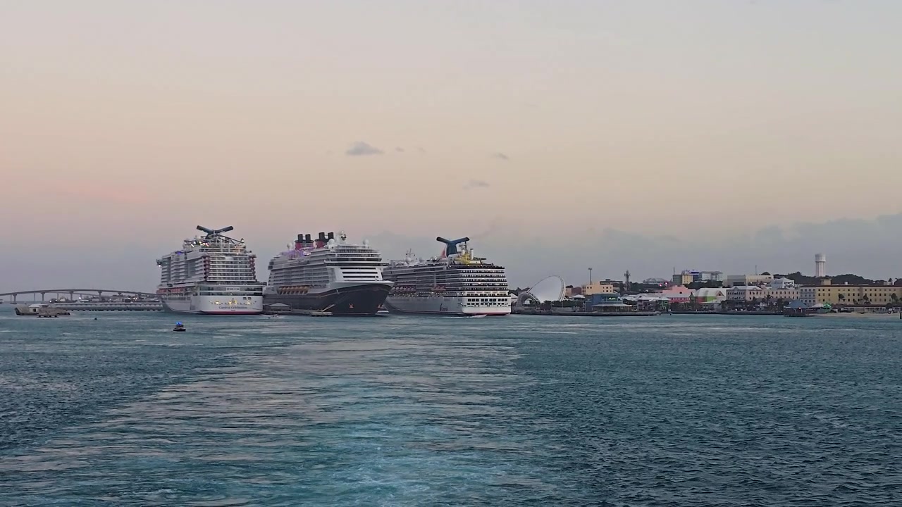 Tre store passagerkrydstogtskibe fortøjet ved en havneterminal i skumringen. Udsigt fra vandet, der viser et kølvand, en kystbys skyline og en bro under en blød aftenhimmel.