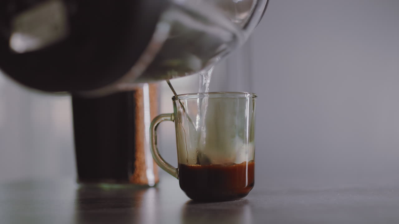 Close up of hot water being poured from kettle into transparent mug containing coffee granules and spoon, steam beginning to rise as drink preparation starts on clean kitchen table surface