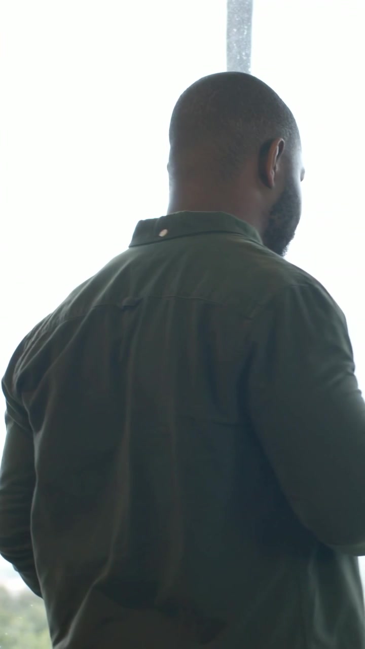 Vertical video: Holding phone, man wearing green shirt, earbud, adjusting blind to see exterior